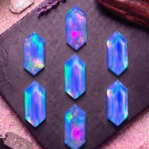 May include: Seven hexagonal, iridescent blue gemstones with internal flashes of pink and green light. Each stone features a faceted, elongated shape. Two small metal tags labeled INDIA GEMS are visible near the corners of the dark display board.