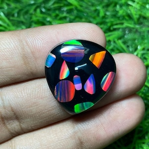 Op de afbeelding: Een zwarte edelsteen met een mozaïek van kleurrijke, iriserende stukken. De gepolijste steen heeft een druppelvorm en wordt in een hand gehouden tegen een achtergrond van groen gras. De kleurrijke stukken omvatten tinten rood, oranje, groen, blauw en paars.