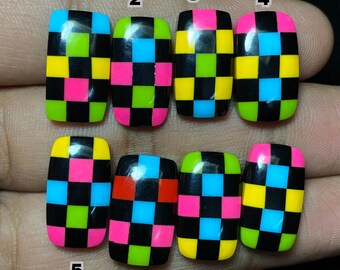 May include: Eight artificial nails with a black checkerboard pattern. Each nail features a unique combination of neon pink, yellow, green, and blue squares. The nails are rectangular with rounded edges, perfect for a bold manicure.
