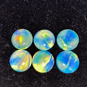 May include: Six round, iridescent cabochons with a polished finish. The gemstones display a mix of blue, yellow, and green hues, with a swirling pattern. The cabochons are set against a black background.