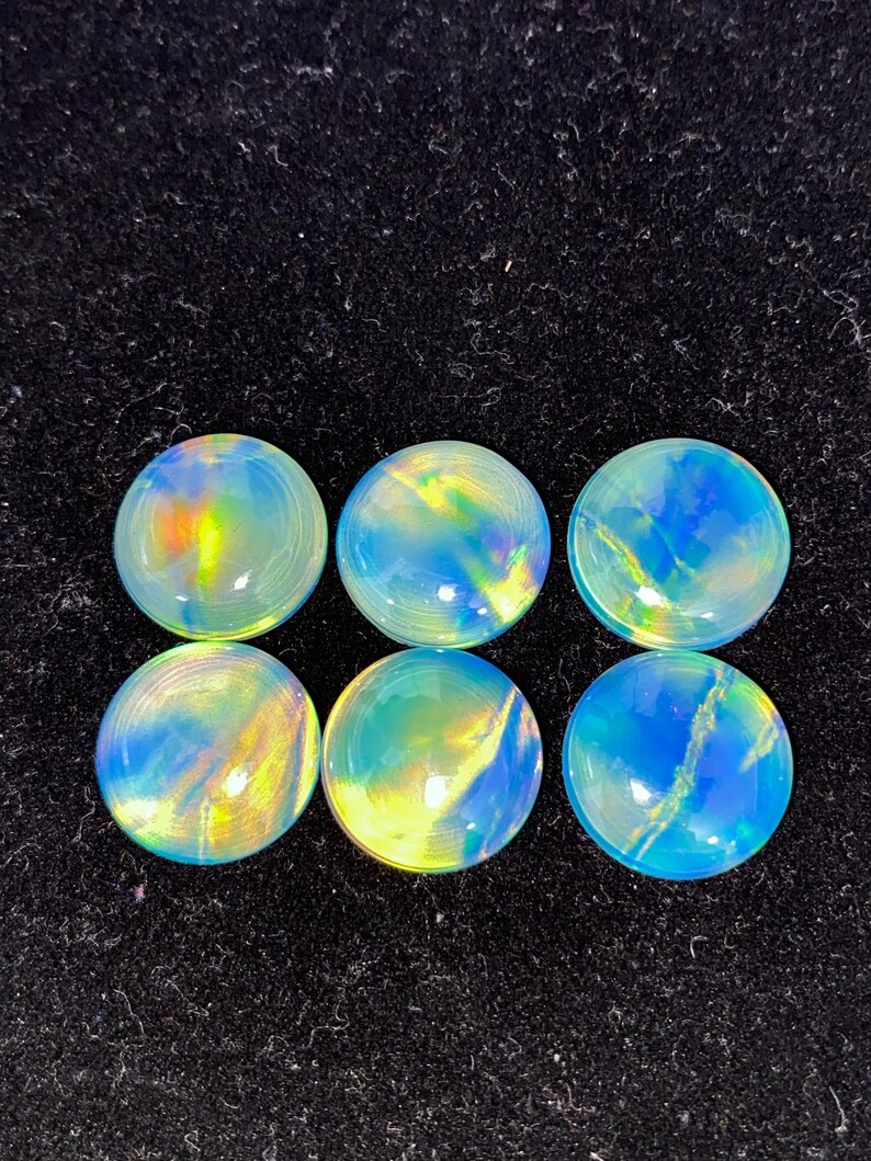 May include: Six round, iridescent cabochons with a polished finish. The gemstones display a mix of blue, yellow, and green hues, with a swirling pattern. The cabochons are set against a black background.
