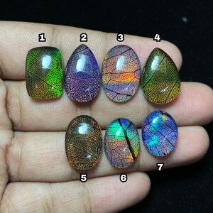 May include: Seven polished gemstone cabochons in various shapes, each displaying a unique leaf-like pattern with vibrant colors. The gemstones feature hues of green, purple, orange, and blue, with intricate veining details. The cabochons are held in a hand against a dark background.