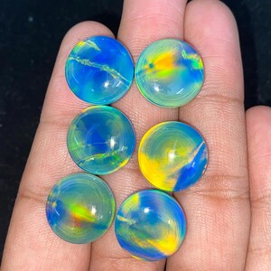 May include: Six round, polished gemstones with vibrant colors of blue, yellow, and green. The stones have a swirling, iridescent appearance, with a glossy finish. The gemstones are held in a hand.