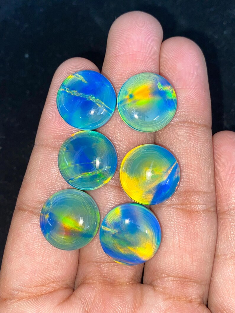 May include: Six round, polished gemstones with vibrant colors of blue, yellow, and green. The stones have a swirling, iridescent appearance, with a glossy finish. The gemstones are held in a hand.