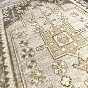 Puede incluir: Una alfombra rectangular con un medallón geométrico central en tonos beige, crema y verde oliva. La alfombra muestra un motivo floral repetitivo en toda la superficie, bordeado por patrones intrincados y rematado con un fleco corto.