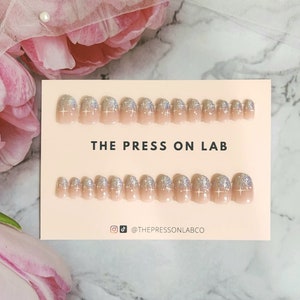 Könnte beinhalten: Ein Set mit 20 Press-on-Nägeln mit einer hellrosa Basis und silbernen Glitzer-Spitzen. Die Nägel sind in zwei Reihen auf einer rosa Karte mit dem Text "THE PRESS ON LAB" und dem Instagram-Handle "@THEPRESSONLABCO" angeordnet.