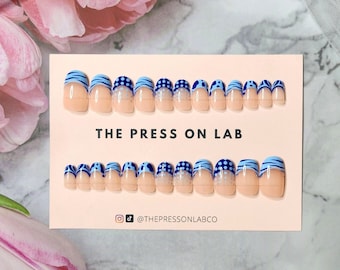 Pode incluir: Um conjunto de 10 unhas postiças com um design azul e branco. As unhas têm uma base rosa claro com um design de listras azuis e brancas, bolinhas azuis e um detalhe branco. As unhas são exibidas em um cartão branco com o texto "THE PRESS ON LAB" e o nome de usuário de mídia social "@THEPRESSONLABCO".