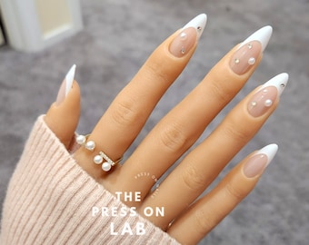 May include: White stiletto press-on nails with a French manicure design and pearl accents. The nails are shown on a hand wearing a gold ring with three pearls.