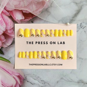 Pode incluir: Um conjunto de 10 unhas postiças com um design de fatia de limão. As unhas são amarelas com uma base branca e têm folhas verdes. As unhas estão dispostas em duas filas em um cartão rosa com o texto "THE PRESS ON LAB" e "THEPRESSONLABLLC.ETSY.COM".