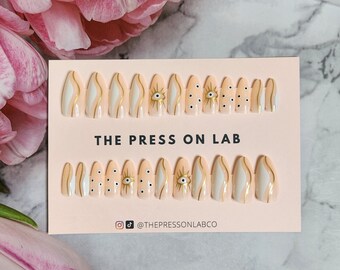 May include: A set of 10 press-on nails with a peachy-pink base and white and gold designs. The nails feature a variety of designs, including eyes, dots, and swirls. The card reads "THE PRESS ON LAB" and includes the social media handle "@THEPRESSONLABCO".
