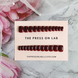 May include: A set of 20 burgundy press-on nails with a gradient effect. The nails are arranged in two rows on a pink card with the text "THE PRESS ON LAB" and "THEPRESSONLABLLC.ETSY.COM".