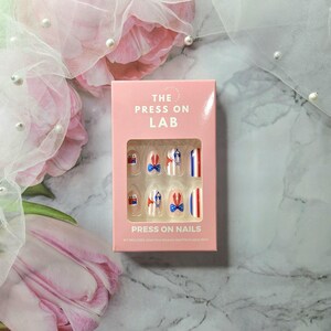 Pode incluir: Uma caixa rosa de unhas posti&ccedil;as com tema patri&oacute;tico. As unhas apresentam desenhos de la&ccedil;os, listras e uma pequena ilustra&ccedil;&atilde;o de um edif&iacute;cio em vermelho, branco e azul. A caixa diz "THE PRESS ON LAB" e "PRESS ON NAILS".