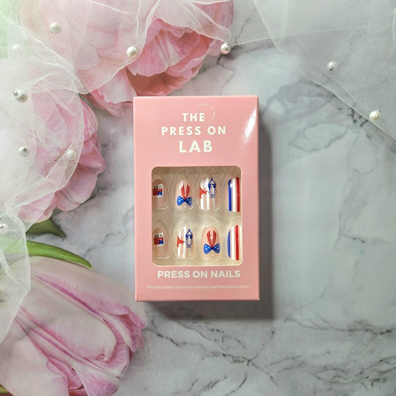 Pode incluir: Uma caixa rosa de unhas posti&ccedil;as com tema patri&oacute;tico. As unhas apresentam desenhos de la&ccedil;os, listras e uma pequena ilustra&ccedil;&atilde;o de um edif&iacute;cio em vermelho, branco e azul. A caixa diz "THE PRESS ON LAB" e "PRESS ON NAILS".