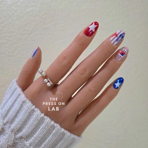Pode incluir: Uma m&atilde;o com unhas posti&ccedil;as com desenhos patri&oacute;ticos em vermelho, branco e azul, incluindo estrelas e listras. Um anel de ouro com p&eacute;rolas e cristais &eacute; vis&iacute;vel. O texto "THE PRESS ON LAB" &eacute; vis&iacute;vel.