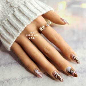 May include: A hand with gingerbread man nail art. The nails are painted a light brown color with white gingerbread man designs. The ring finger has a white and red candy cane design. The hand is wearing a ring with three white pearls. The text "THE PRESS ON LAB" is visible on the hand.