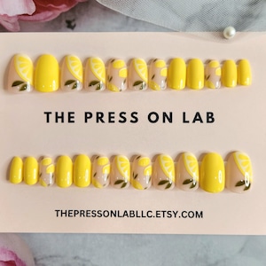 May include: Two sets of press-on nails. Each set has a variety of sizes. Some nails are solid yellow, while others feature a lemon slice design with green leaves on a nude background. The text "THE PRESS ON LAB" is visible.