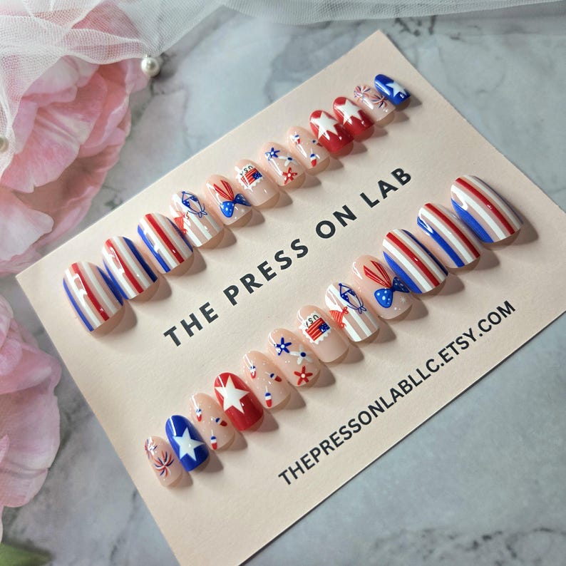 Pode incluir: Uma cole&ccedil;&atilde;o de unhas posti&ccedil;as com designs patri&oacute;ticos. As unhas apresentam listras vermelhas, brancas e azuis, estrelas e outros gr&aacute;ficos festivos. O texto "THE PRESS ON LAB" e "THEPRESSONLABLLC.ETSY.COM" est&aacute; impresso no cart&atilde;o.