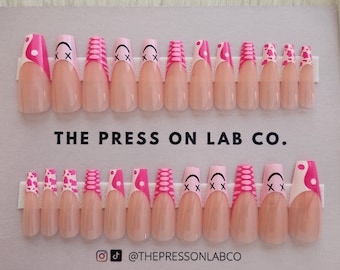 Puede incluir: Un conjunto de 20 uñas postizas de color rosa y blanco con diferentes diseños, incluyendo caras sonrientes, X y lunares. Las uñas están dispuestas en dos filas sobre un fondo gris claro. El texto "THE PRESS ON LAB CO." está impreso debajo de las uñas. El texto "@THEPRESSONLABCO" está impreso debajo del texto.