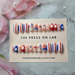 Pode incluir: Um conjunto de unhas posti&ccedil;as com tema patri&oacute;tico. As unhas apresentam listras vermelhas, brancas e azuis, estrelas e desenhos de la&ccedil;os. O texto "THE PRESS ON LAB" e "THEPRESSONLABLLC.ETSY.COM" s&atilde;o vis&iacute;veis.