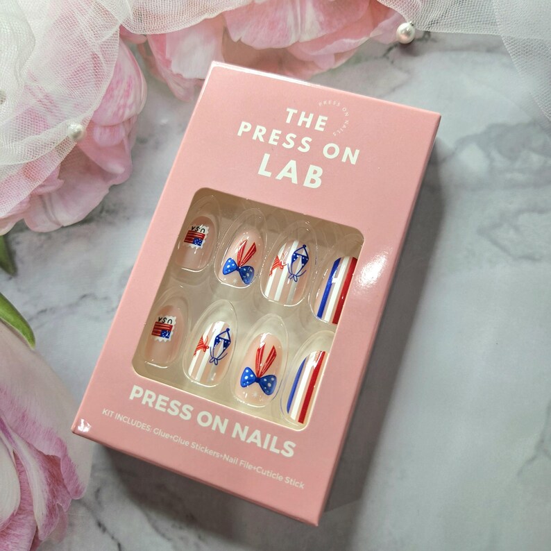 Pode incluir: Uma caixa rosa de unhas posti&ccedil;as com designs patri&oacute;ticos. As unhas apresentam listras, la&ccedil;os e gr&aacute;ficos da bandeira dos EUA em vermelho, branco e azul. A caixa diz "THE PRESS ON LAB" e "PRESS ON NAILS".