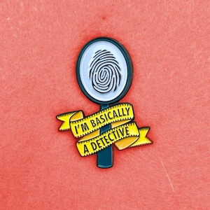 Può includere: Spilla a tema detective con una lente d'ingrandimento che mostra un'impronta digitale. Un banner giallo sottostante recita "I'M BASICALLY A DETECTIVE". Lo sfondo è rosso.