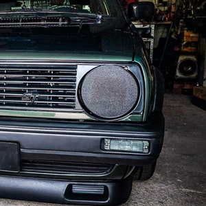 Op de afbeelding: Een groene Volkswagen Golf GTI met een zwarte voorbumper en een zwarte grille. De auto heeft een zwart en zilveren GT-badge op de grille. De auto staat in een garage.
