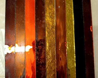 May include: A collection of rectangular glass pieces in various colors, including shades of brown, orange, gold, and black. The pieces are arranged side by side, with a ruler placed below them for scale. The glass has a glossy, reflective surface.