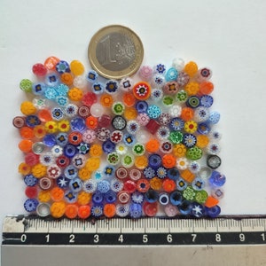 May include: A selection of vibrant millefiori glass beads, featuring floral and geometric designs. The beads display a range of colours, including blue, orange, yellow, red, and green. A Euro coin is placed nearby for scale. A ruler is at the bottom.