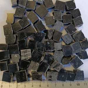 Puede incluir: Un conjunto de pequeñas baldosas cuadradas de color gris oscuro, posiblemente de piedra o cerámica, dispersas sobre una superficie. Las baldosas varían ligeramente en tono y textura, algunas mostrando patrones sutiles. Una regla es visible en la parte inferior.