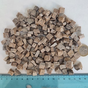 May include: A pile of small, irregular, light brown and beige stone tiles. The tiles are about 1/2 inch in size and have a rough, textured surface. A ruler is visible in the bottom of the image.