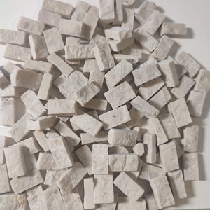 Puede incluir: Un montón de pequeñas baldosas rectangulares de piedra de color blanco roto. Las baldosas miden aproximadamente 2,5 cm de largo y 1,25 cm de ancho. Se puede ver una regla en la parte inferior de la imagen, con marcas del 1 al 15.