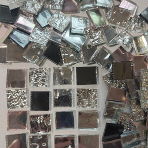May include: A collection of small, square, iridescent glass tiles. The tiles are arranged in a grid pattern, with some tiles having a textured surface. The tiles are a mix of silver, black, and iridescent colors.