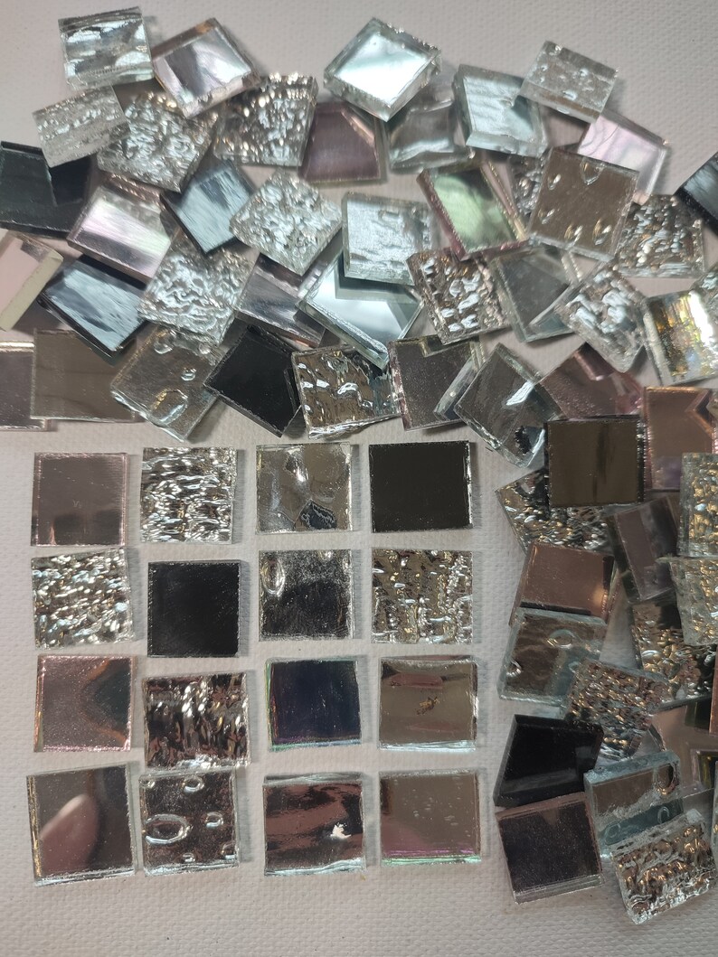 May include: A collection of small, square, iridescent glass tiles. The tiles are arranged in a grid pattern, with some tiles having a textured surface. The tiles are a mix of silver, black, and iridescent colors.