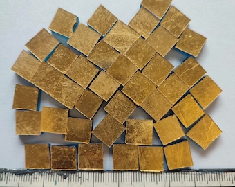 May include: A pile of small, square gold leaf tiles with a blue border. The tiles are scattered on a white surface with a ruler measuring 9 inches.