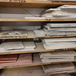 May include: A wooden shelf unit with various types of marble tiles stacked on each shelf. The tiles are labeled with names like "Nembro Rosato", "Travertine Classico", "Breccia Paradiso", "Botticino", "Travertine Rosso", "Travertine Noce", and "Darnosio".
