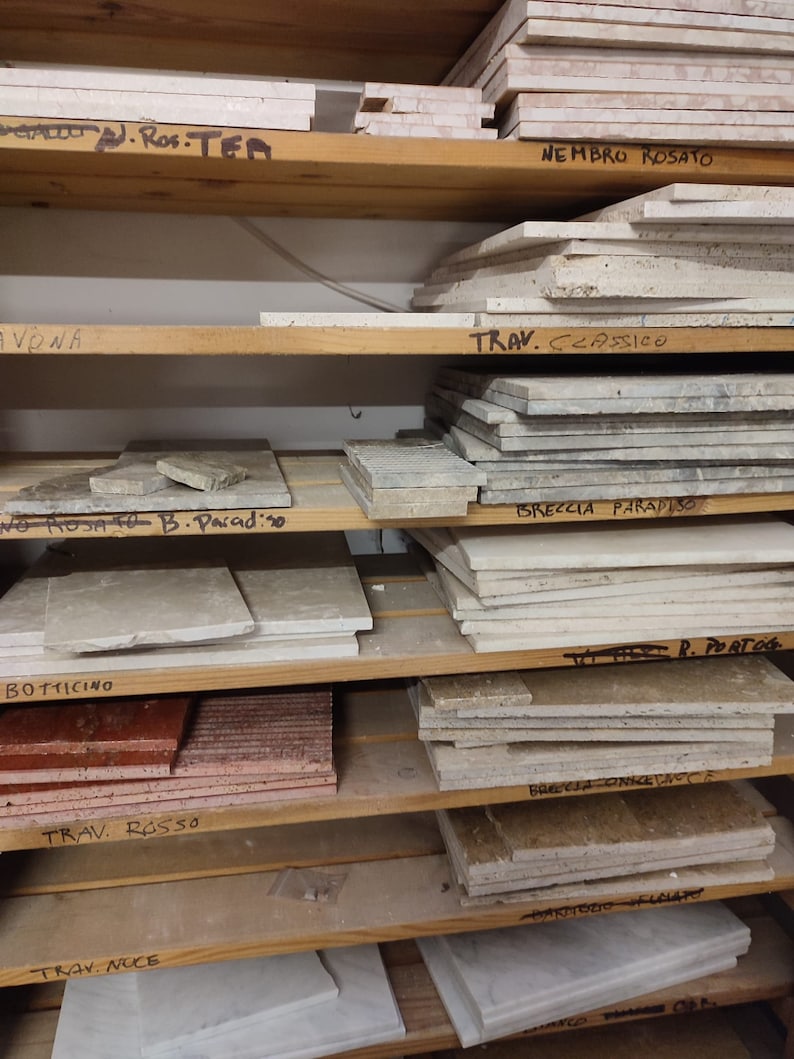 May include: A wooden shelf unit with various types of marble tiles stacked on each shelf. The tiles are labeled with names like "Nembro Rosato", "Travertine Classico", "Breccia Paradiso", "Botticino", "Travertine Rosso", "Travertine Noce", and "Darnosio".