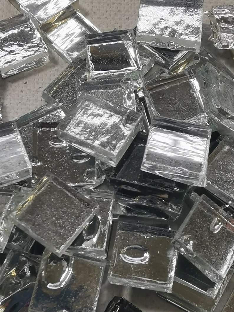May include: A pile of small, clear glass tiles with a textured surface. The tiles are square and have a slightly iridescent sheen.