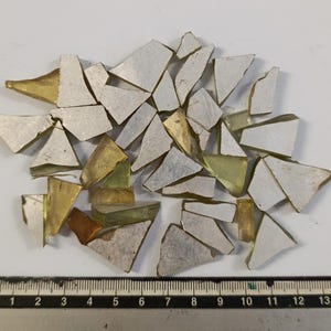 May include: A collection of irregularly shaped glass pieces with silver and gold tones. The fragments are scattered across a white surface, with a ruler at the bottom for scale. The pieces vary in size and shape, creating a mosaic effect.