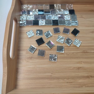 May include: A wooden tray with a mosaic tile design. The tiles are black, silver, and pink. Some of the tiles are loose and scattered around the tray.