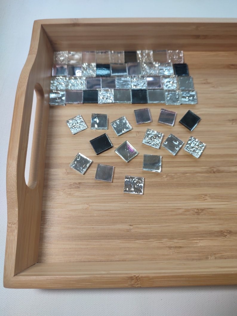 May include: A wooden tray with a mosaic tile design. The tiles are black, silver, and pink. Some of the tiles are loose and scattered around the tray.