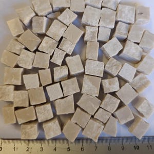 Puede incluir: Un conjunto de pequeñas teselas de mosaico beige, de forma cuadrada. Las teselas están dispersas, con tamaños variables y texturas ligeramente rugosas. Una regla es visible en la parte inferior, indicando medidas en centímetros. Adecuado para manualidades.