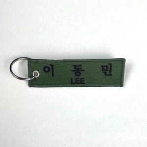 May include: A green fabric keychain with black embroidered text. The keychain has a silver metal ring attached to one end. The text includes Korean characters and the English name "LEE".