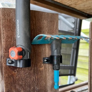 May include: Two gardening tools are mounted on a wooden wall. A black and red Gardena tool is on the left and a blue rake is on the right. Both tools are attached to the wall with black metal brackets.
