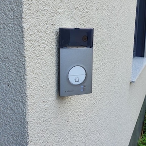 Op de afbeelding: Een zilver en zwart deurbelsysteem gemonteerd op een getextureerde, lichtbeige buitenmuur. De deurbel heeft een witte knop met een belpictogram, een kleine camera en indicatielampjes. De merknaam is zichtbaar.