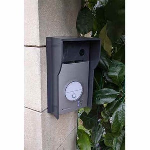 Może przedstawiać: Szary dzwonek do drzwi z białym przyciskiem i małym obiektywem kamery. Dzwonek jest zamontowany na kamiennej ścianie.