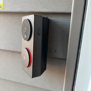 Op de afbeelding: Een zwarte deurbelcamera met een ronde, zilveren knop en een lens met een zwarte ring eromheen. De camera is gemonteerd op een grijze muur.