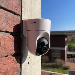 Może przedstawiać: Biała kamera bezpieczeństwa zamontowana na ceglanej ścianie. Kamera ma czarny obiektyw i białą obudowę. Kamera jest skierowana w stronę ulicy.