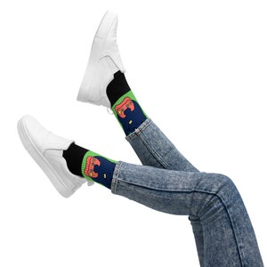 May include: A pair of white sneakers with black socks featuring a green and red gamepad design. The person is wearing blue jeans.