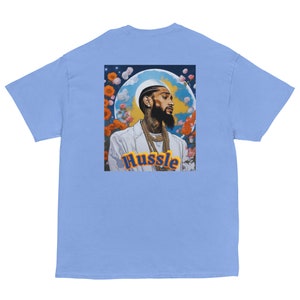 May include: A light blue t-shirt with a graphic print of a man with a beard and a crown, surrounded by flowers and clouds. The text "Hustle" is printed in yellow and blue letters below the image.