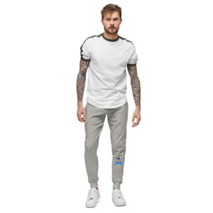 May include: A man is wearing a white t-shirt with a green trim and gray sweatpants with a blue and yellow graphic on the left leg.